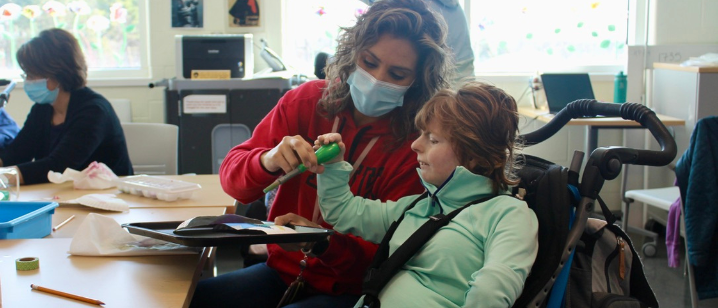 A teaching assistant helps a student in a wheelchair hold a paint brush.