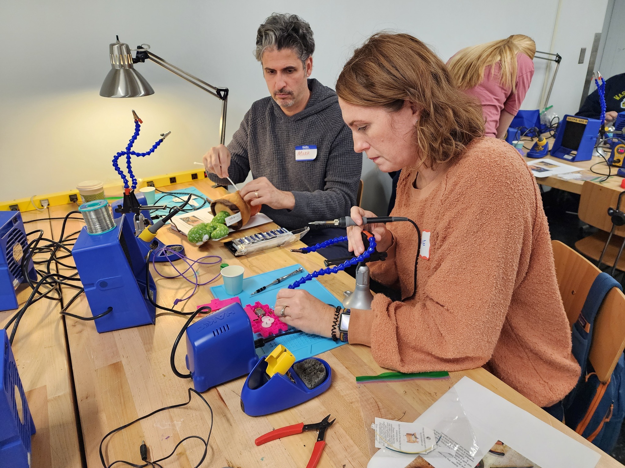 Two individuals sit at a table and modify toys using various tools